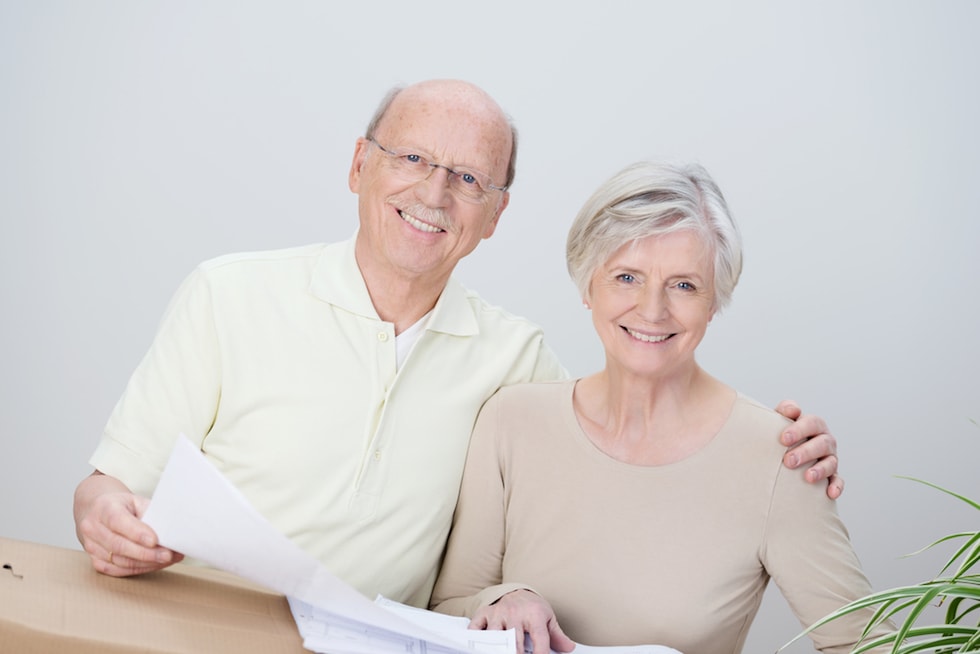 Smiling affectionate senior couple standing with the husbands arm around his wife looking at documents resting on a brown cardboard carton as they plan their move to a new home-2